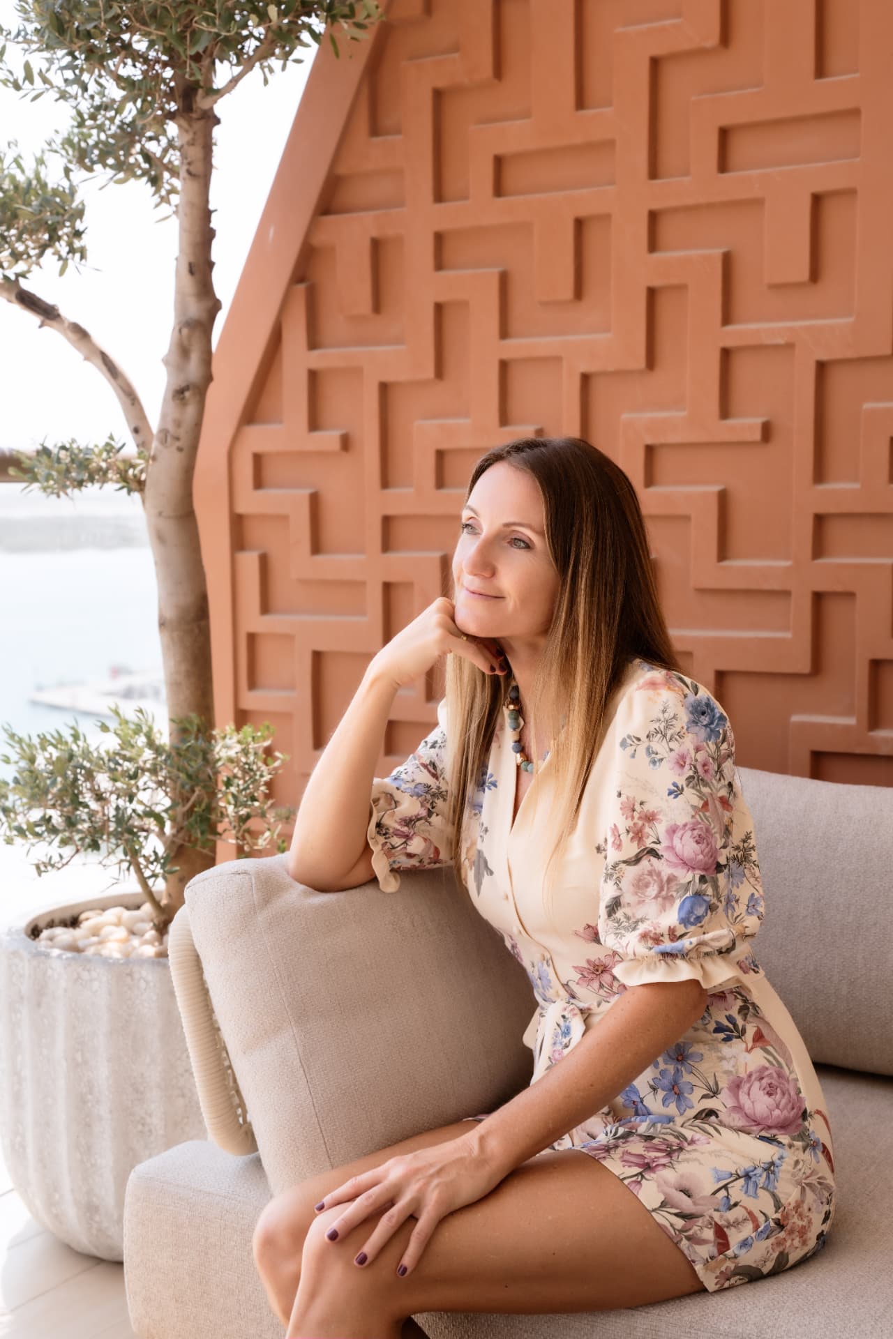 Andrea Mathis seated thoughtfully on a terrace sofa in a floral dress, beside an olive tree and a terracotta patterned wall